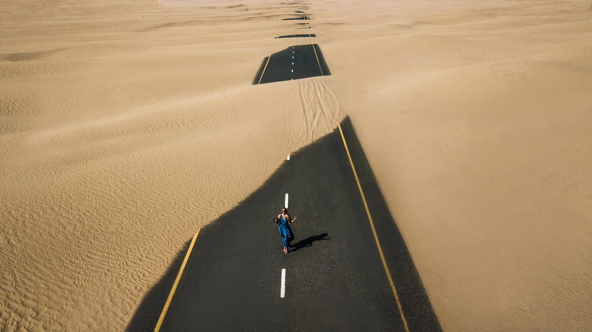 Aerial view of woman in blue dress walking along desert road with yellow lines, representing the journey from desert hike to SAUSSAVITA's Velvet Rose fragrance.