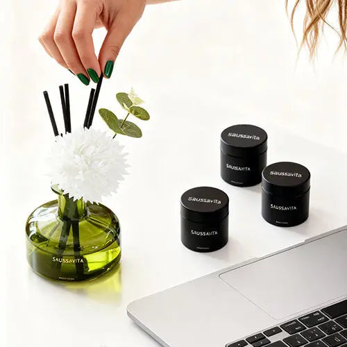 Green-nailed hand places reed into SAUSSAVITA diffuser near black containers and laptop - home fragrance setup.
